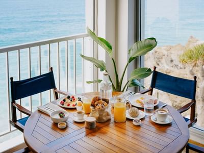 A table with a seaside breakfast, featuring croissants, juices, and coffee, set by a balcony overlooking the ocean.