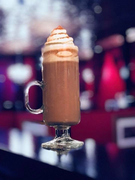 A tall glass mug of hot chocolate sitting on a bar, topped with whipped cream and cocoa powder, found at Better than Sex in Orlando.