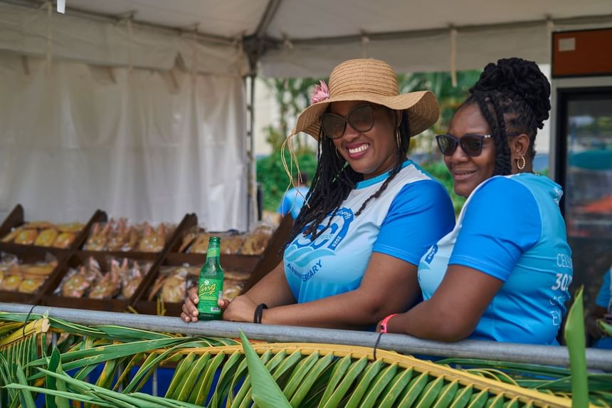 Two women stand behind a bamboo barrier with snacks at Taste The Bay 2025 near Bay Gardens Hotels and Resorts