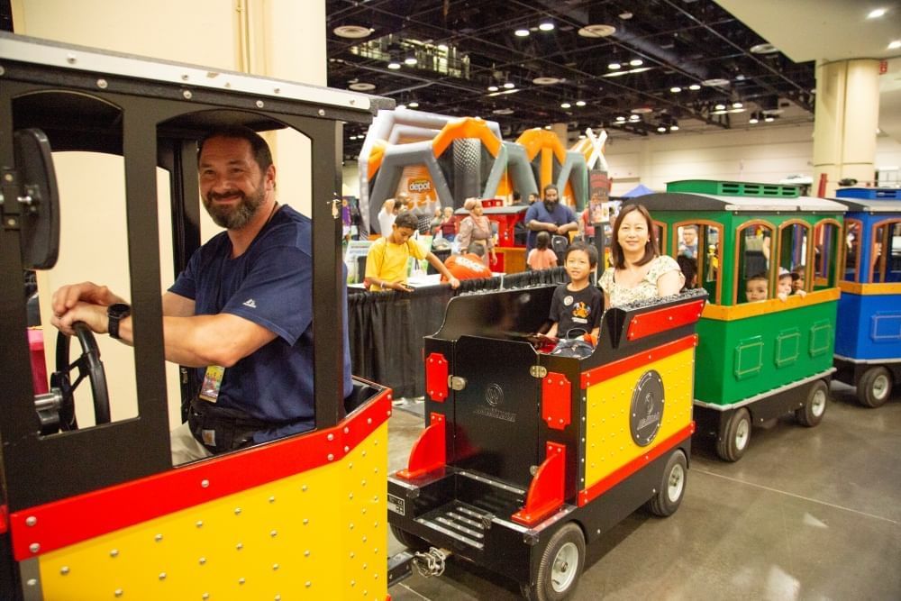 A man drives a small, colorful train with children and parents as passengers through an indoor expo.
