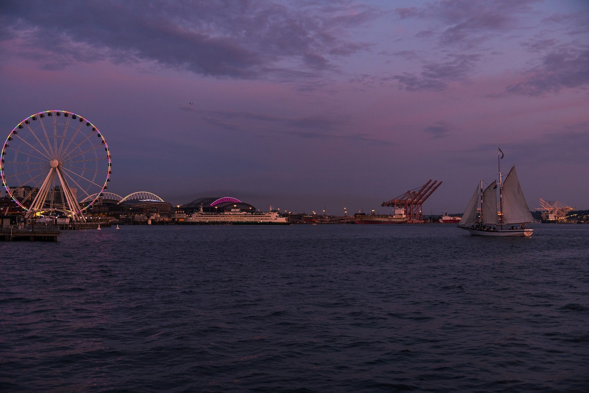 a wide view of a waterfront area with a sailboat, pier, ferris wheel, and city skyline at sunset