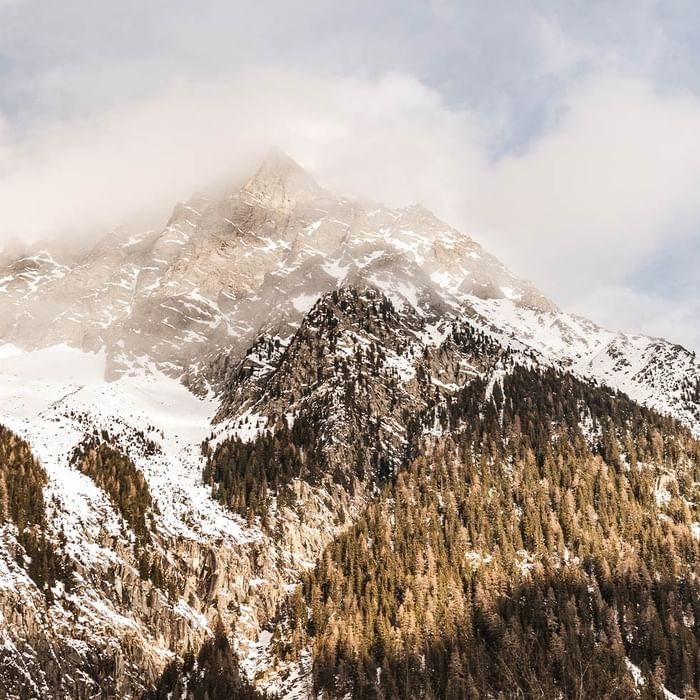 Snowy mountain peaks with clouds and dense forest of evergreen trees at the base.