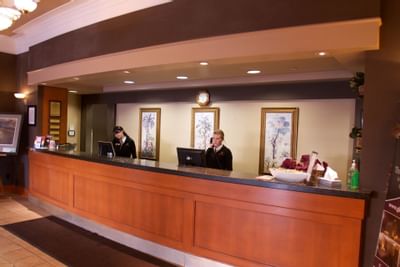 Front desk staff by a wooden counter, framed botanical art under a wall clock at Glenmore Inn & Convention Centre