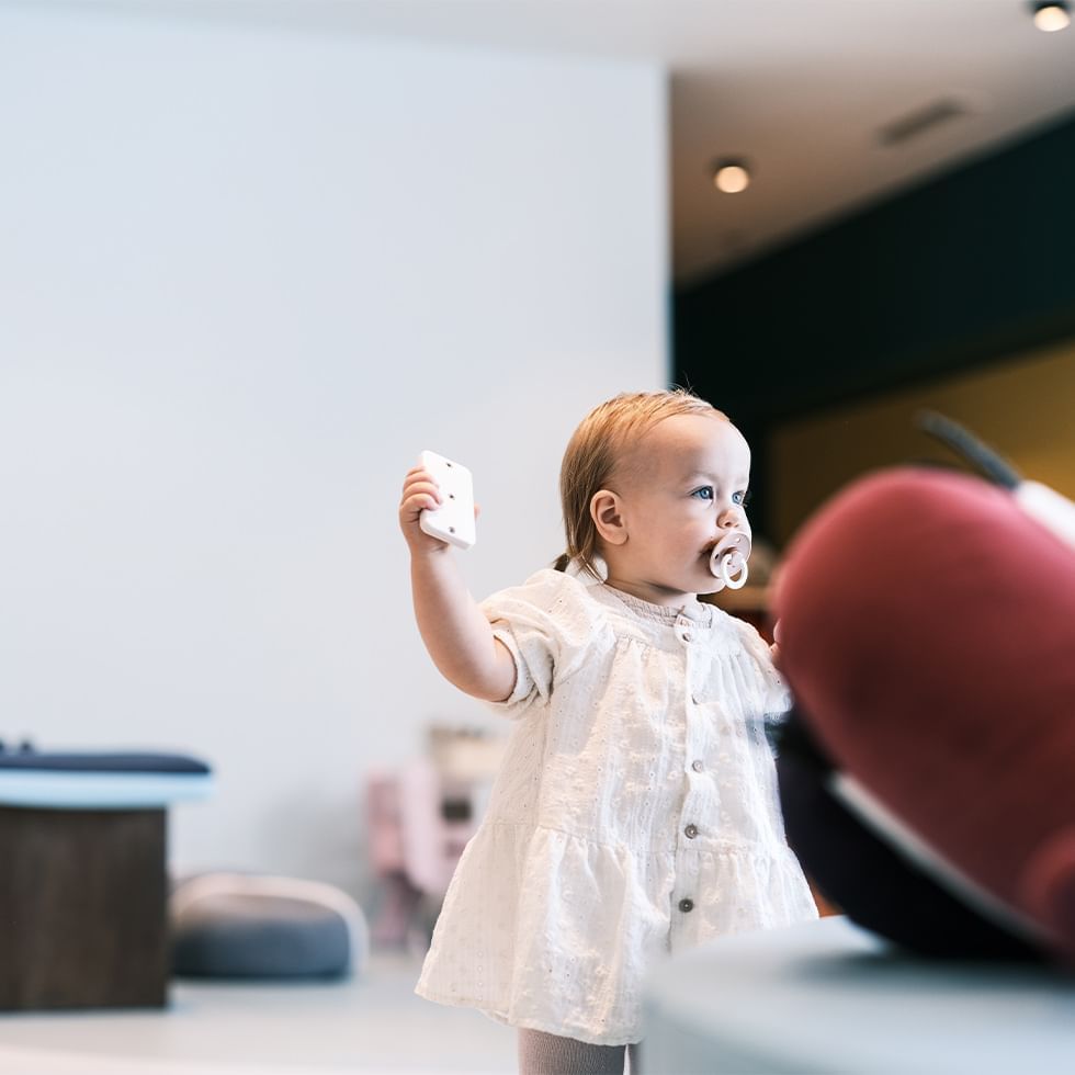 A baby with a pacifier waves a toy in a playroom highlighting Baby Birthday Experience.