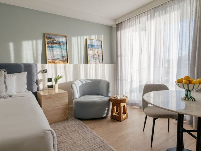 Sunset View Deluxe King at Elysium Manly featuring a marble table by a grey armchair under sunlit curtains near a bed