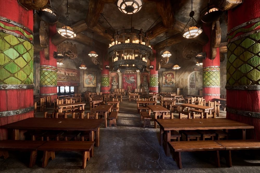 A rustic looking dining hall with wooden picnic tables, hanging wooden chandeliers, and large red pillars with green wooden scales. 
