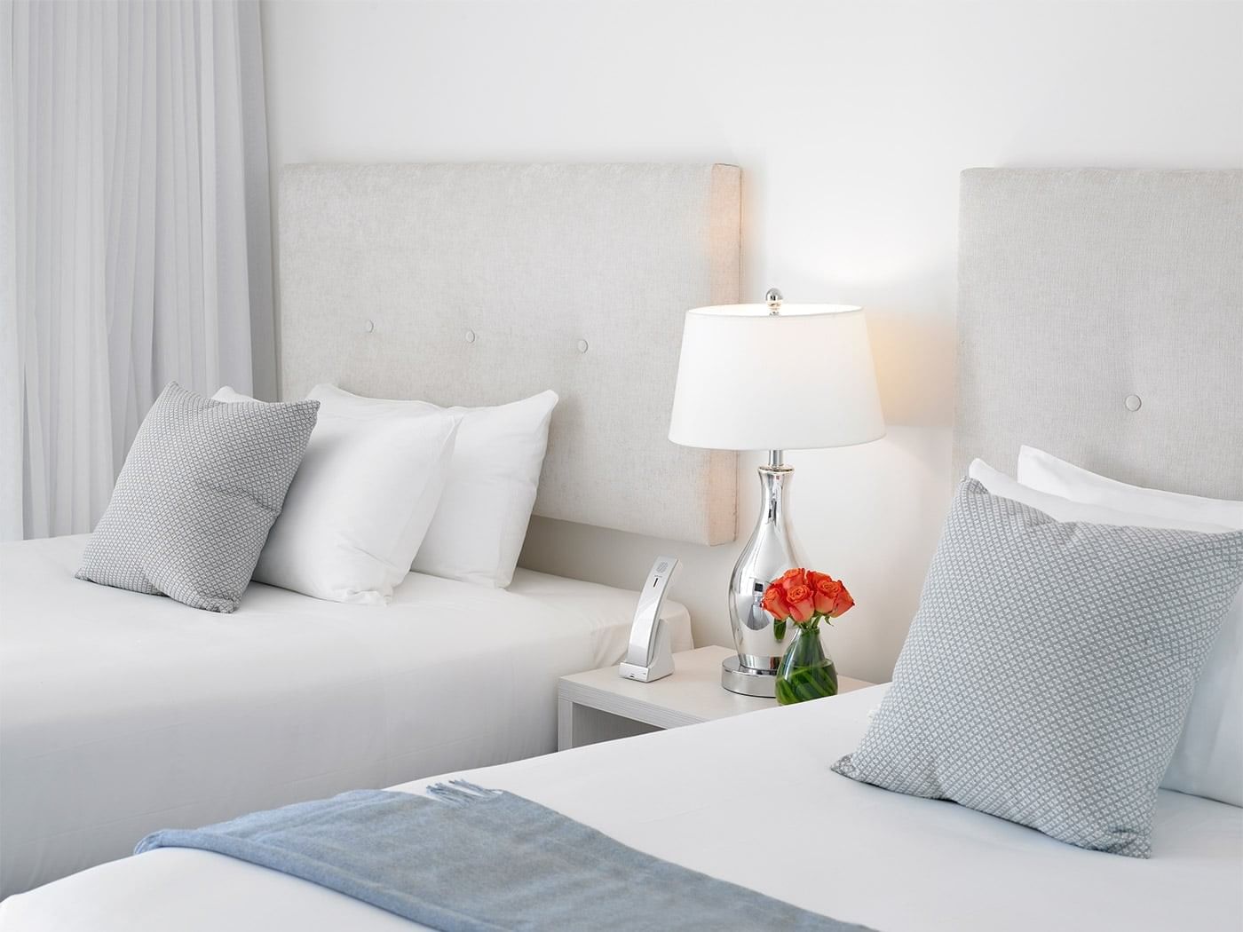 A close-up of a neatly made bed with white sheets, two gray pillows, and a gray blanket at the foot, with a nightstand and lamp in the background.