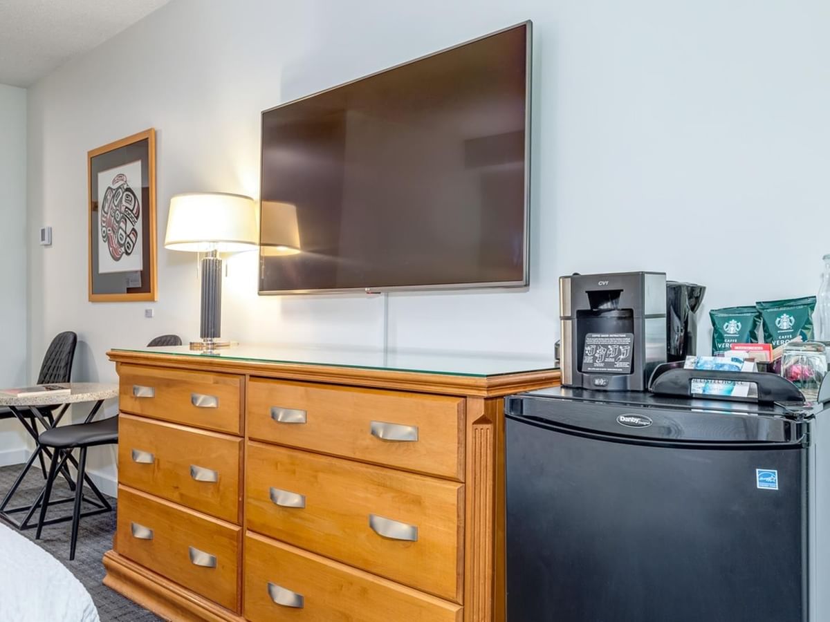 A room with a bed, dresser, refrigerator, coffee maker, lamp, and a TV mounted on the wall.