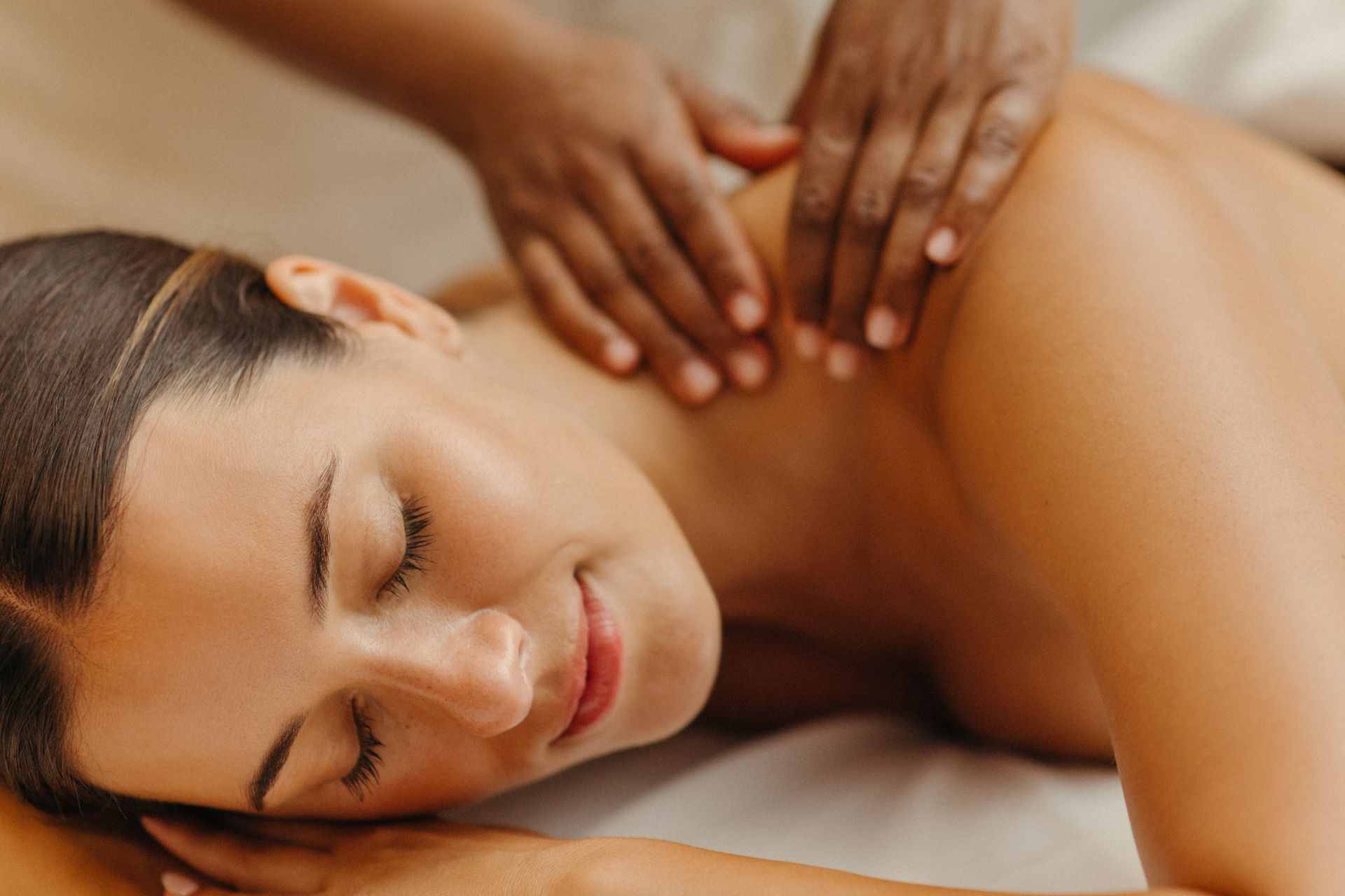 A woman is receiving a shoulder massage, lying down with eyes closed and smiling.