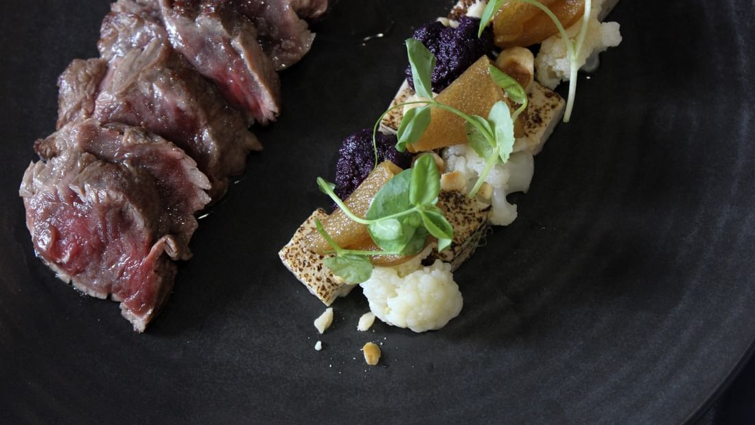 A close-up view of a gourmet dish with meat, vegetables, and garnishes on a black plate.
