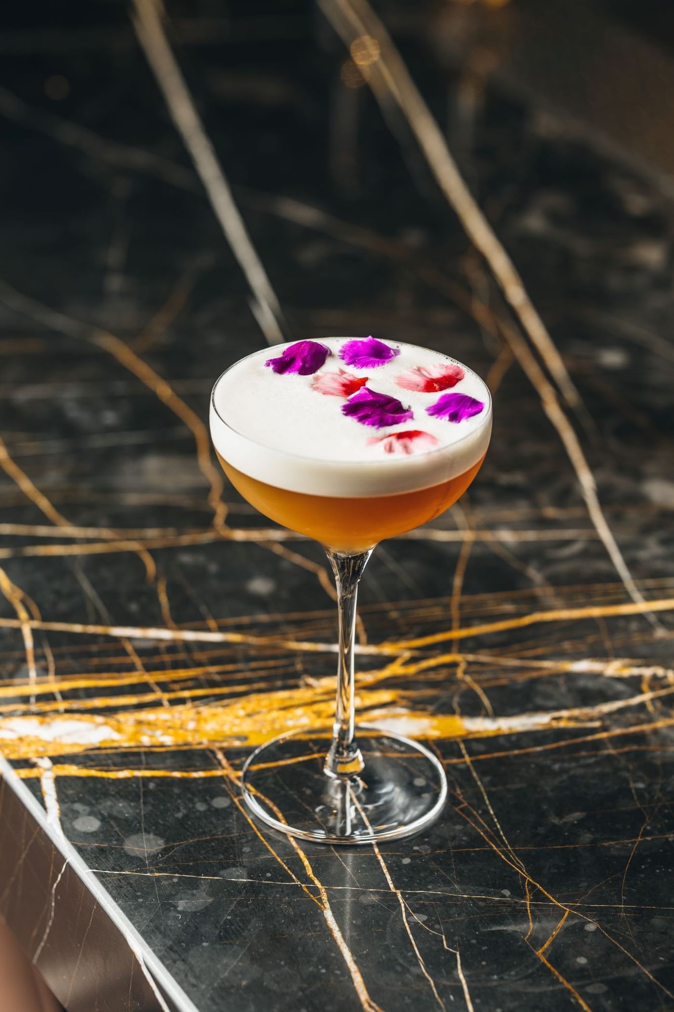 An elegant cocktail garnished with colorful flower petals sits on a polished black marble surface at The Londoner Hotel