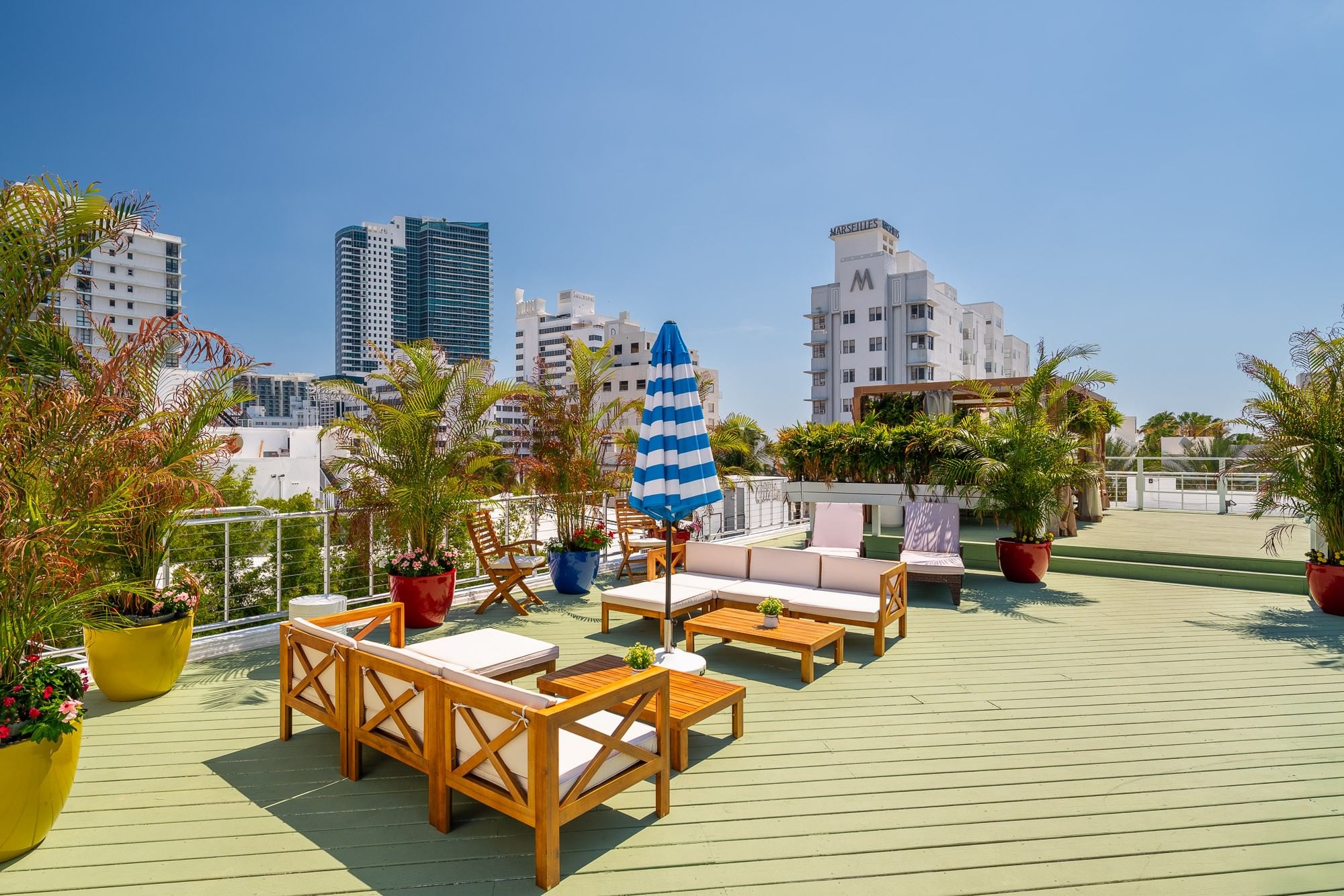 A rooftop deck with green flooring, wooden furniture, potted plants, and a blue and white umbrella, overlooking the city.