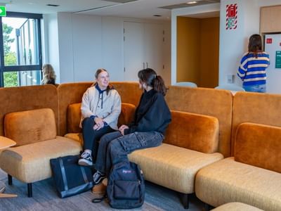 Two guests conversing on orange modular seating in a bright common area with windows and informational displays at Tupuārangi