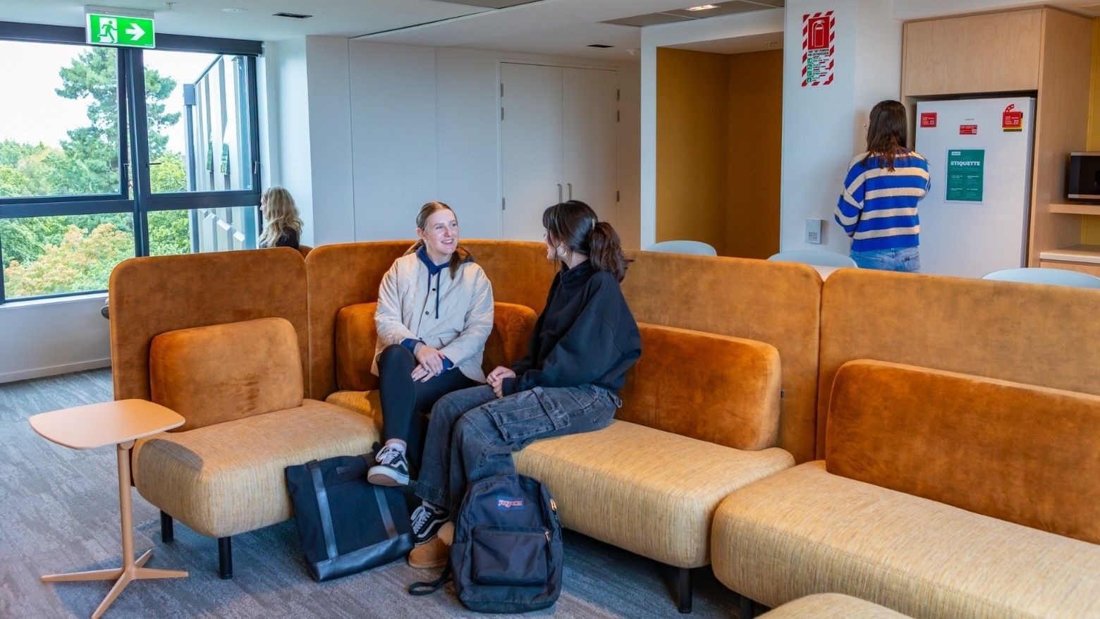 Two guests conversing on orange modular seating in a bright common area with windows and informational displays at Tupuārangi