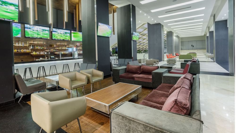 Armchairs by a wooden table under TVs near the illuminated bar counter in Stock Bar at Real Inn Tijuana