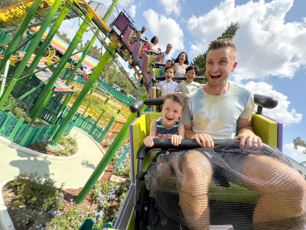 A man and a boy with protective mesh covering their laps ride a roller coaster car and smile as they zoom along a green coaster track. 