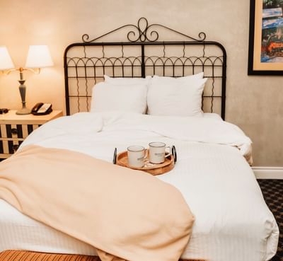 A neatly made bed with white bedding, two mugs, and a throw blanket, alongside a nightstand and framed artwork.
