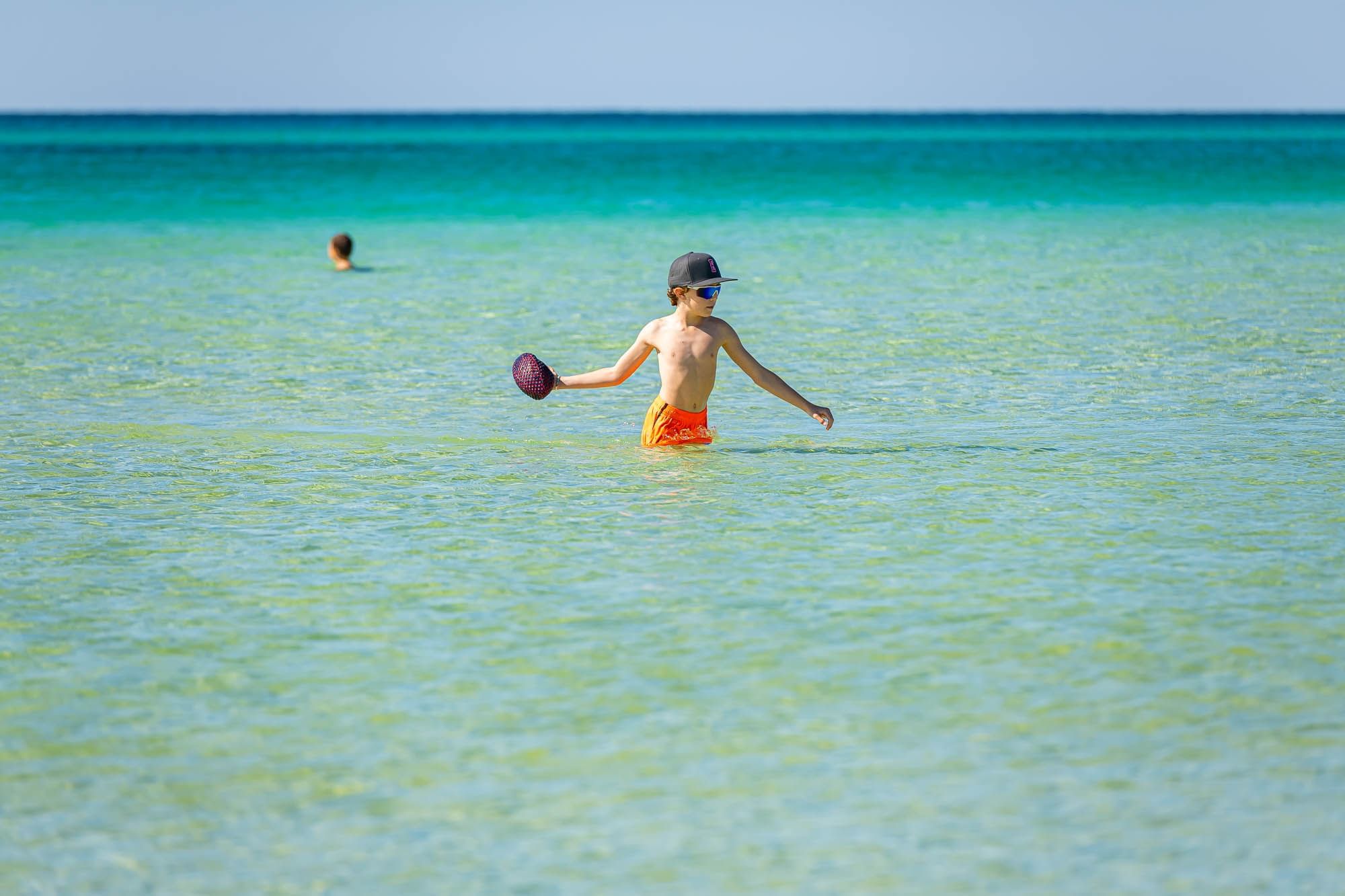 Boy in orange swim trunks and cap holding a frisbee while standing in shallow water.