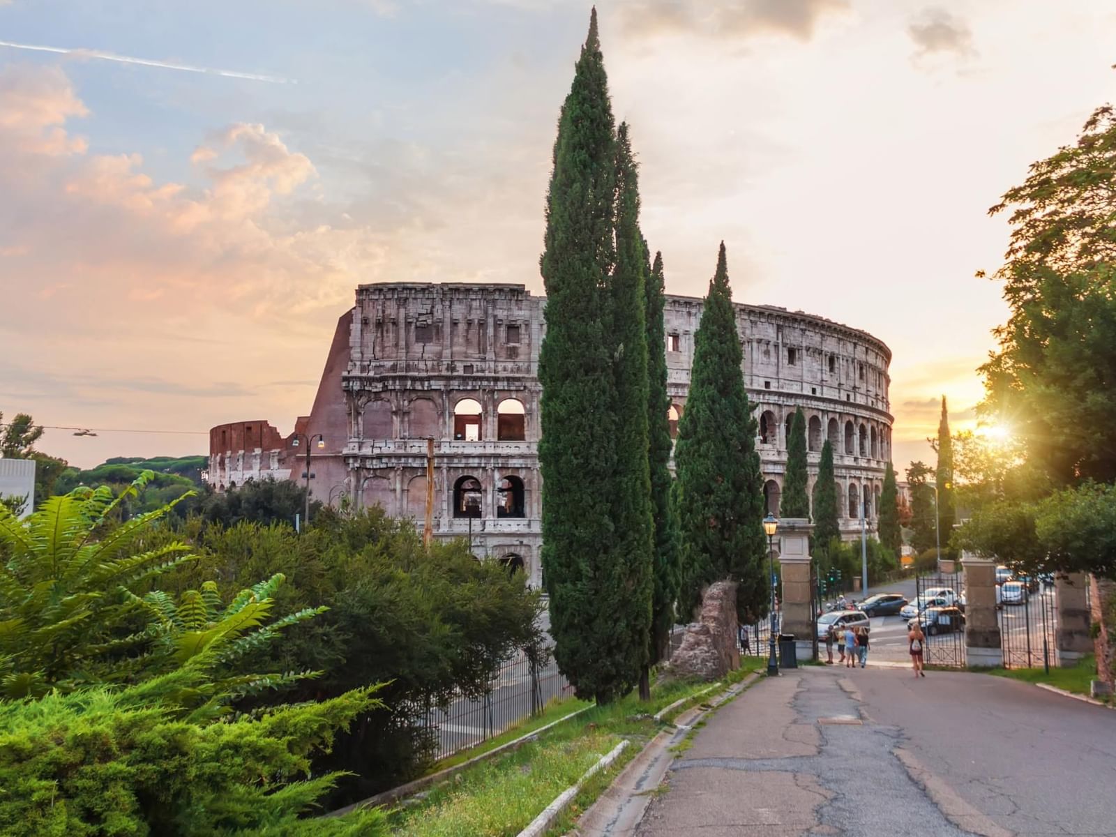 Colosseum at sunset, framed by cypress trees as visitors stroll a quiet street near The Glam Hotel