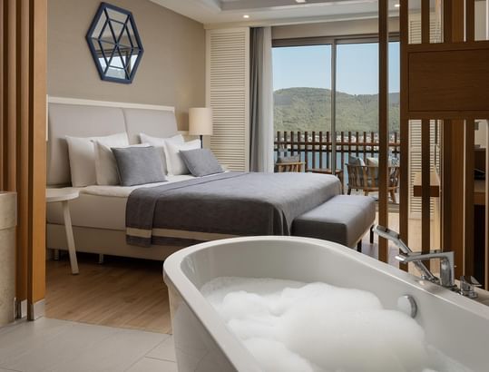 Elegant bathroom with large bathtub, wooden dividers, and bedroom view with balcony and mountain scenery at Titanic Luxury Collection Bodrum.