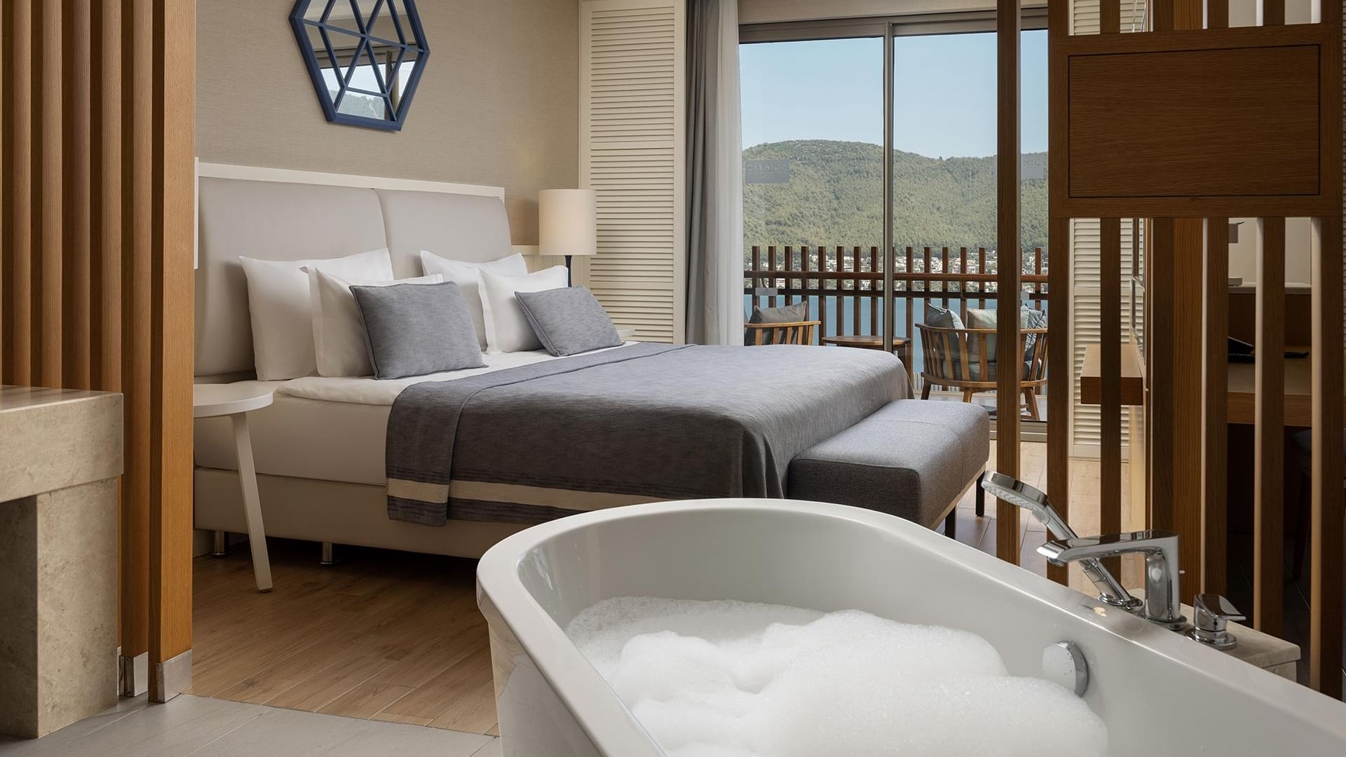 Elegant bathroom with large bathtub, wooden dividers, and bedroom view with balcony and mountain scenery at Titanic Luxury Collection Bodrum.