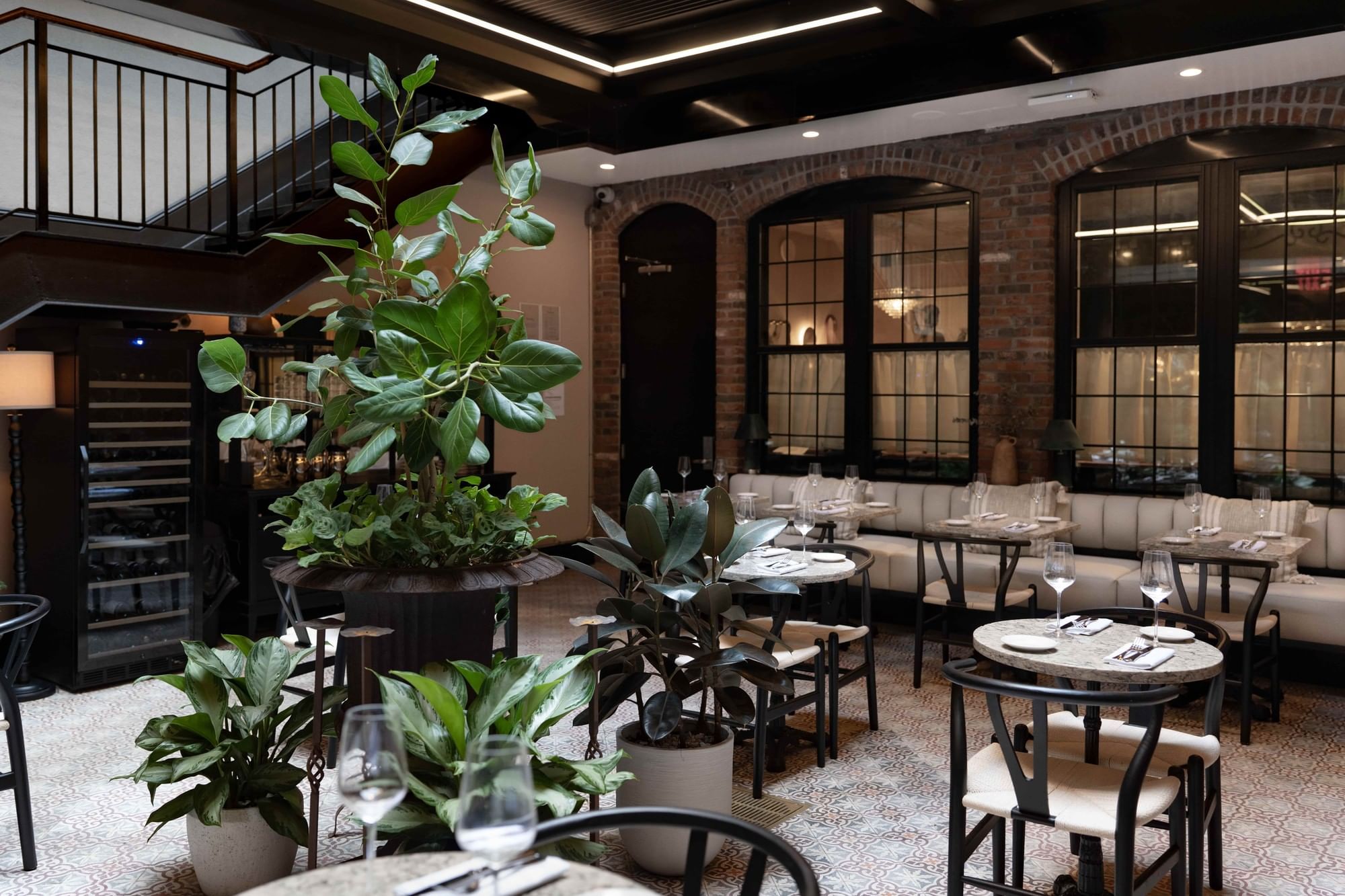 A dining area with tables and chairs, plants, and a brick wall with windows.