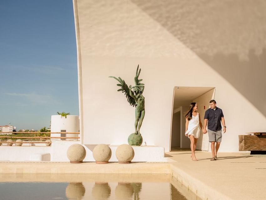 A couple holding hands walks past a large statue and reflective pool at Marquis Los Cabos Resort under blue skies