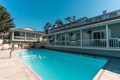 The sunny outdoor swimming pool surrounded by a stone patio and the light blue hotel buildings at Carmel Bay View Inn