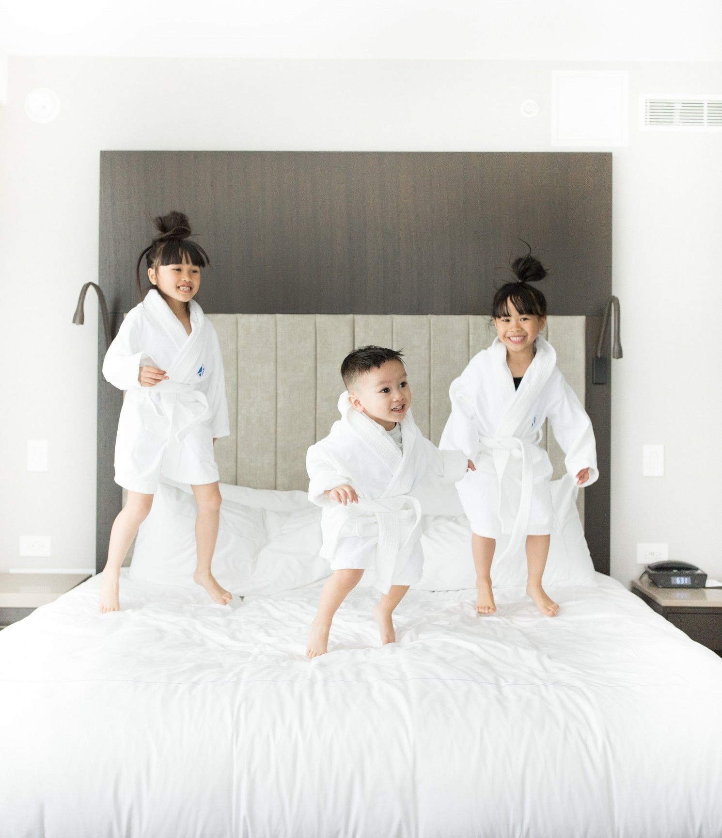 Three children wearing white bathrobes jumping happily on a plush king bed in a room at Hotel X Toronto