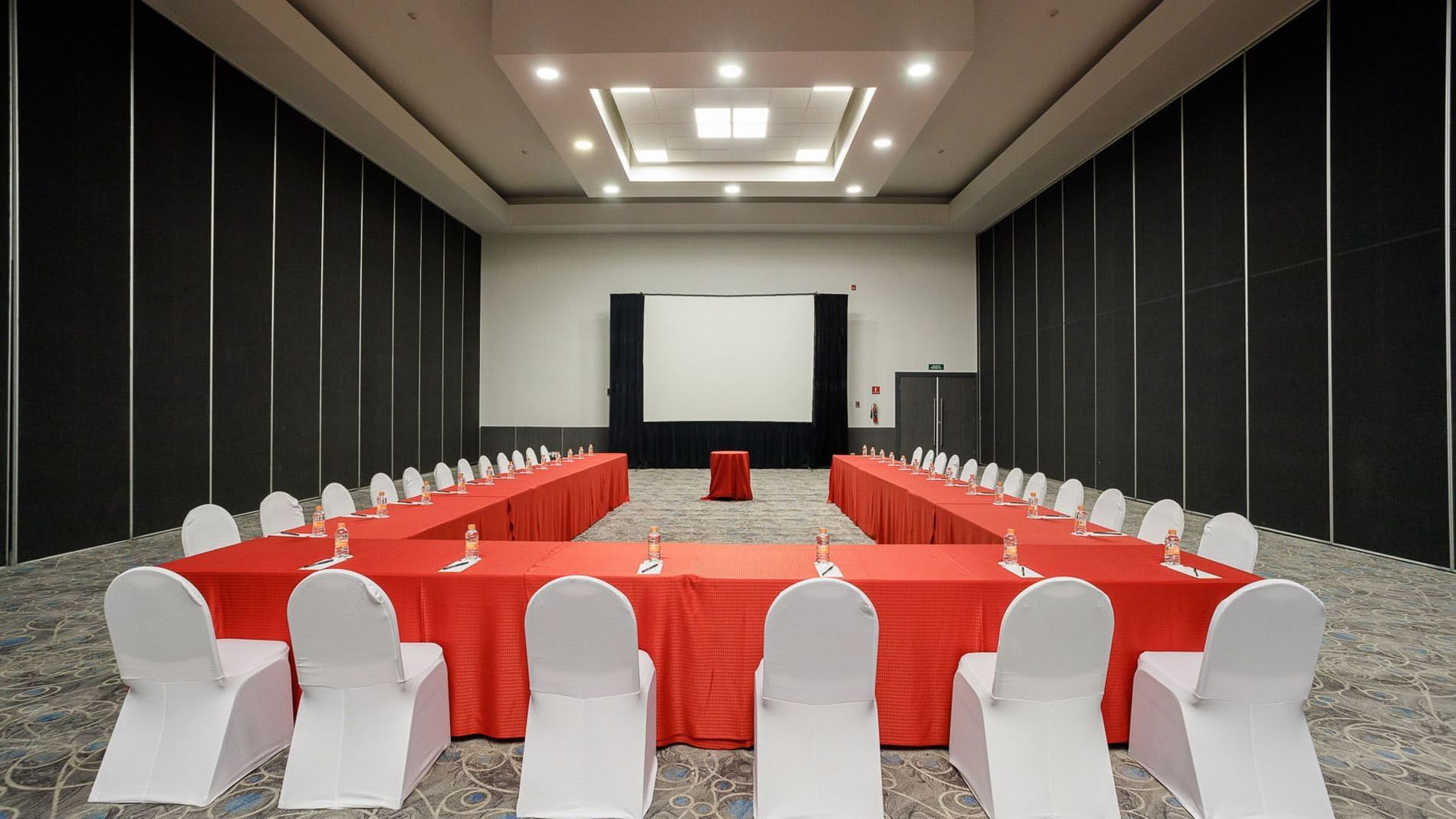 U-shaped meeting setup with red tablecloths and a large projector screen in Venecia Venue at Real Inn Tijuana