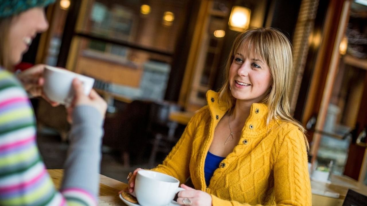 Woman in yellow sweater smiling while enjoying coffee with a friend in a warm, inviting cafe setting.