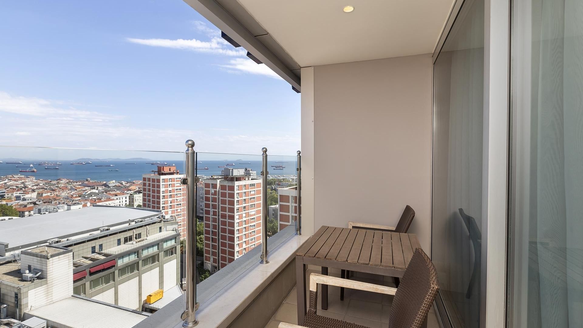 An indoor balcony with a glass wall, a table and chairs, overlooking a stunning cityscape and the ocean beneath a clear blue sky in Executive Room at Titanic Port Bakırköy