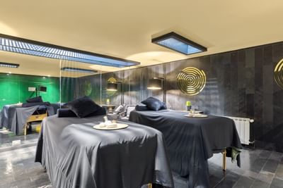 Dos camas de masaje en salón de spa con paredes verdes y negras, iluminación dorada y accesorios de lujo.