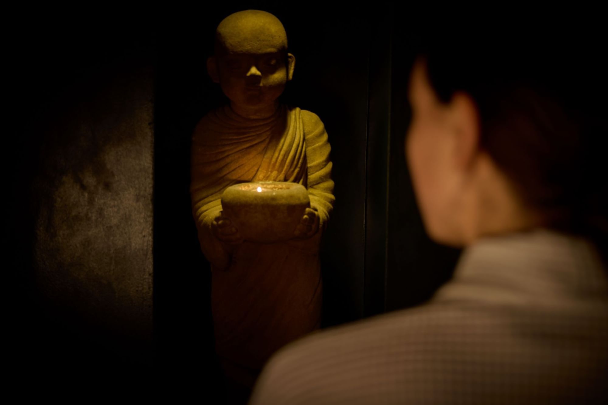 Person looking at a lit candle held by a buddha statue in a dark room at the May Fair Spa.