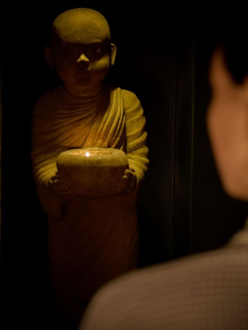 Person looking at a lit candle held by a buddha statue in a dark room at the May Fair Spa.