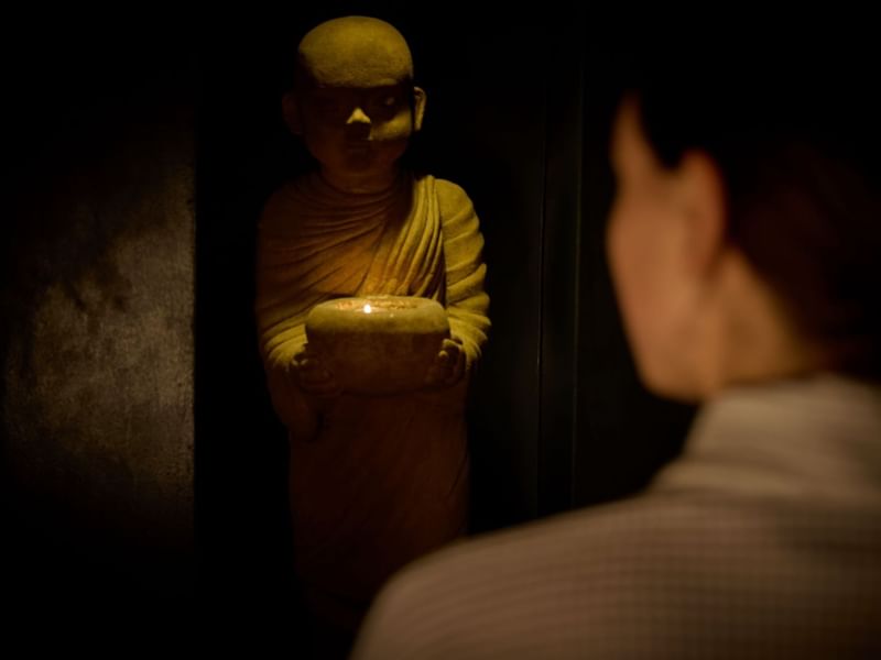 Person looking at a lit candle held by a buddha statue in a dark room at the May Fair Spa.