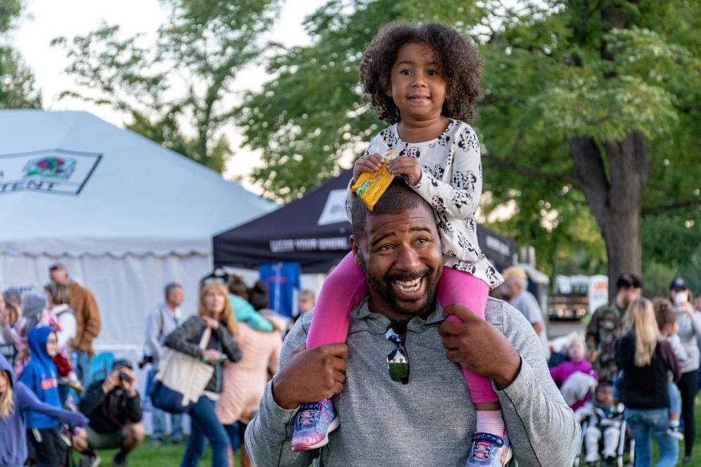 A man laughs carrying a girl on his shoulders at an outdoor festival with tents. 