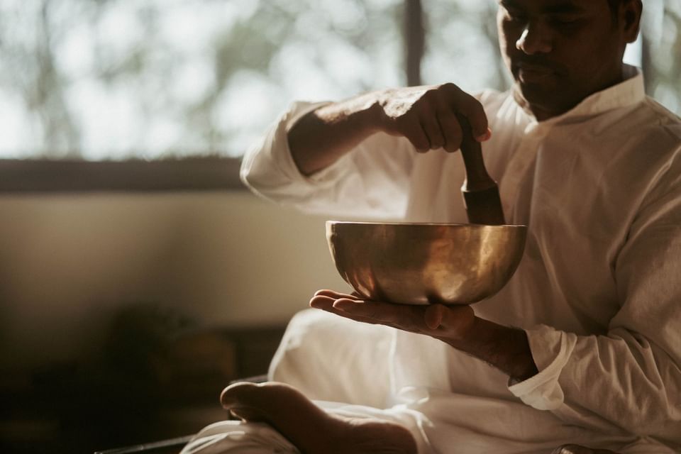 Person in a white robe holds a singing bowl, using a mallet to create soothing sounds at The Dwarika’s Hotels and Resorts