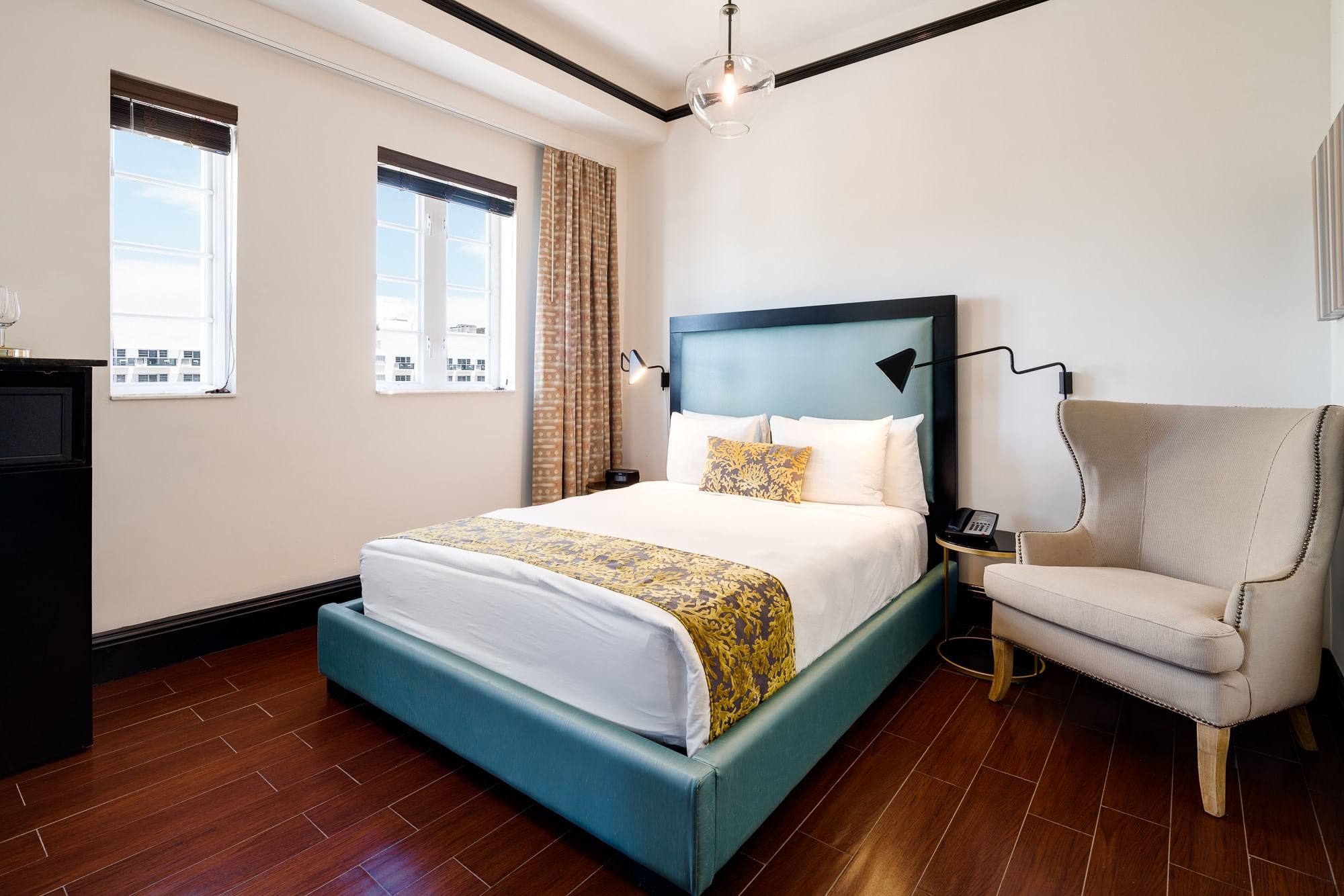 A neatly made king bed with a teal velvet headboard and footboard, accompanied by a beige armchair and a window with a city view.