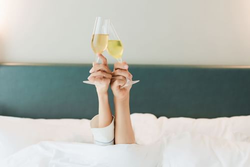 Two hands holding champagne glasses while lying in bed at Warwick Hotels and Resorts