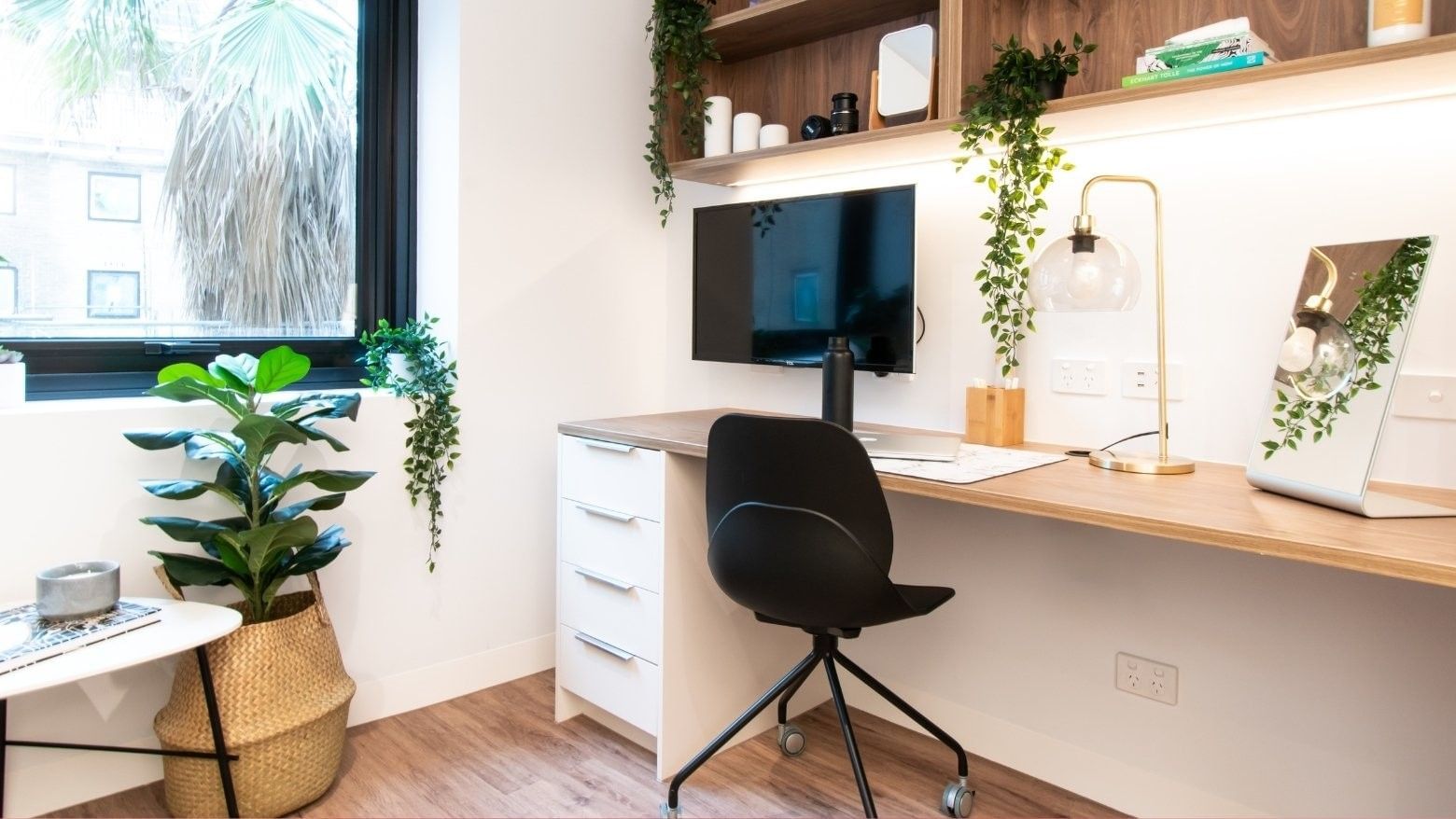 A stylish office space with a desk, chair, monitor, lamp, potted plants, and a shelf with items and a framed picture.