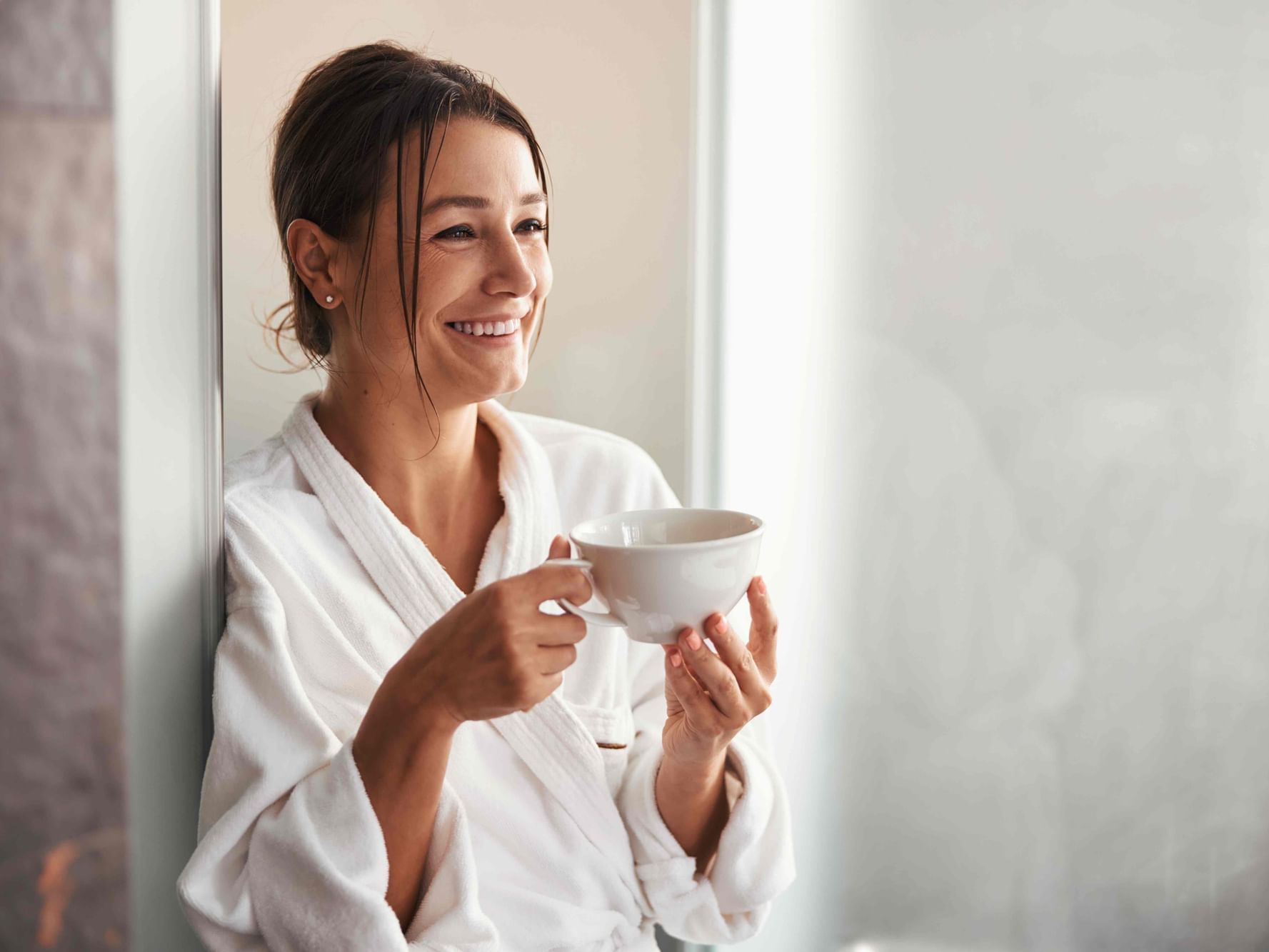A smiling woman in a white hotel bathrobe holding a cup of coffee while looking out a window in a bright hotel room at Hotel Grand Chancellor Townsville.