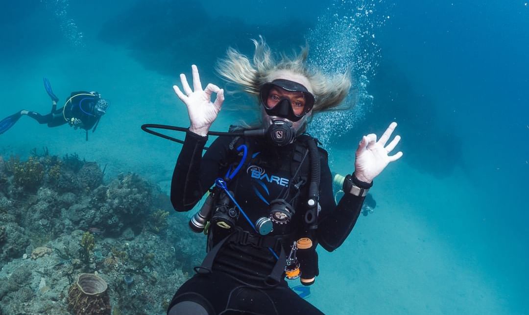 Scubaduiker poseert onder water, maakt een handgebaar, omringd door levendig koraal nabij Golden Rock Resort