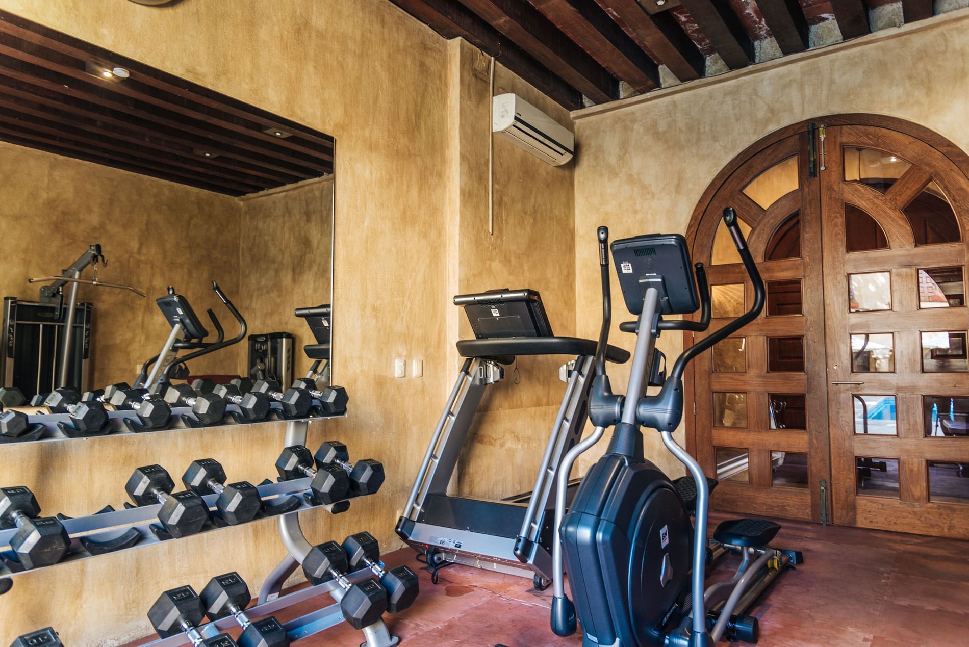 Modern gym with warm-toned walls, featuring a treadmill, elliptical, and a rack of dumbbells at Quinta Real Oaxaca