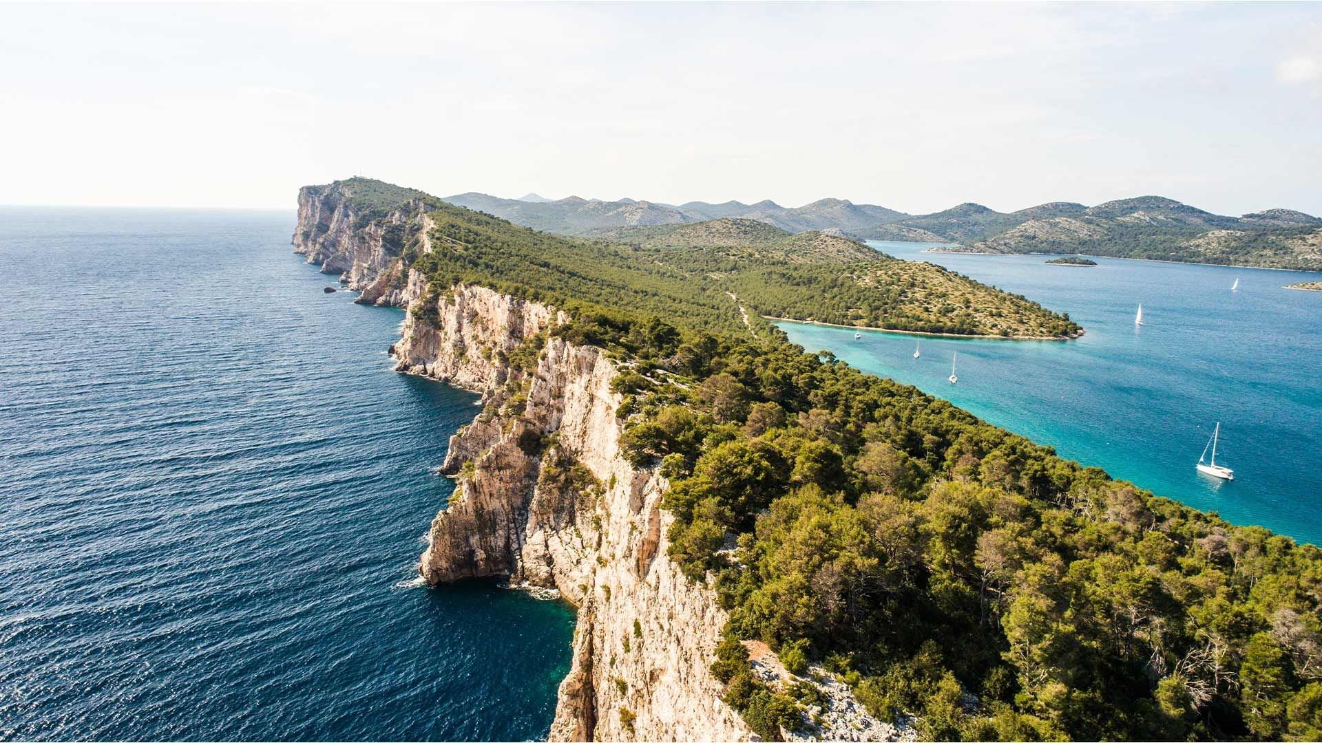 Luftaufnahme einer felsigen Küste mit blauem Meer, bewaldeten Hügeln und Segelbooten im Hintergrund.