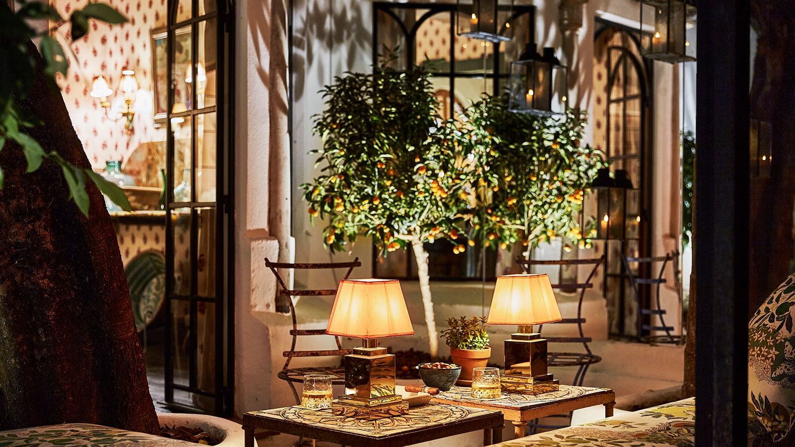 Two glowing brass lamps placed on side tables near a kumquat tree on a dimly lit outdoor terrace at Marbella Club