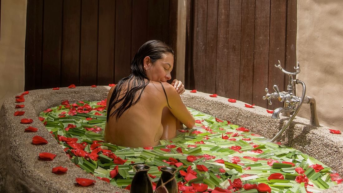 Woman relaxing in a stone bathtub filled with rose petals and green leaves promoting spa treatment and long stay package