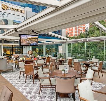 An outdoor seating area with wooden tables, cushioned chairs, and a canopy, offering a view of the trees in Titanic Coffee at Titanic Port Bakırköy