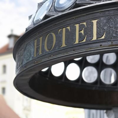Metal sign with "HOTEL" in gold letters, ornate design, and partial view of a building behind.