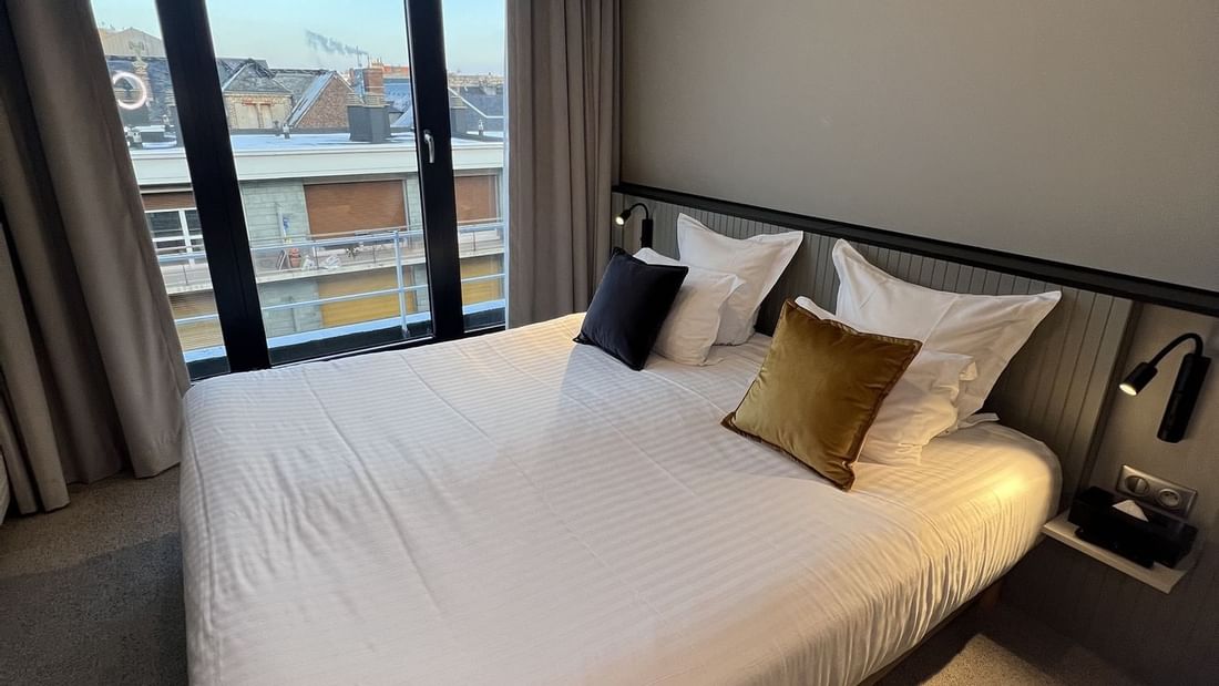 Modern bedroom with large bed, white linens, multiple pillows, and cityscape view through large windows.